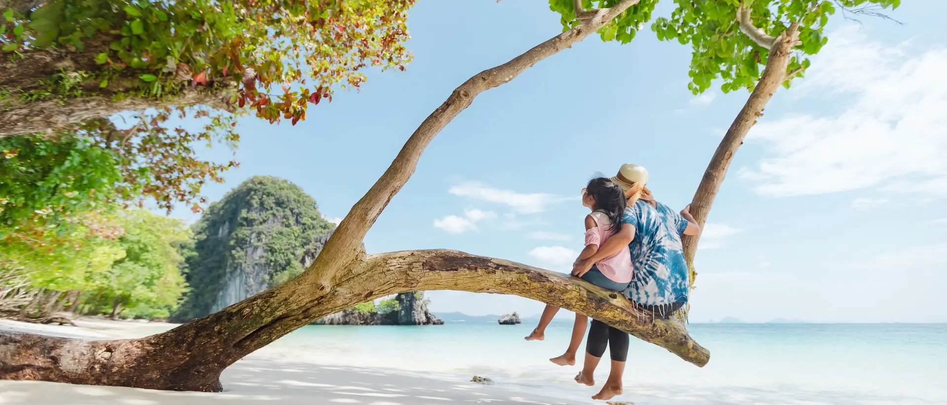 A woman sits on a tree branch by the beach, holding a child, with clear water and lush greenery in the background.