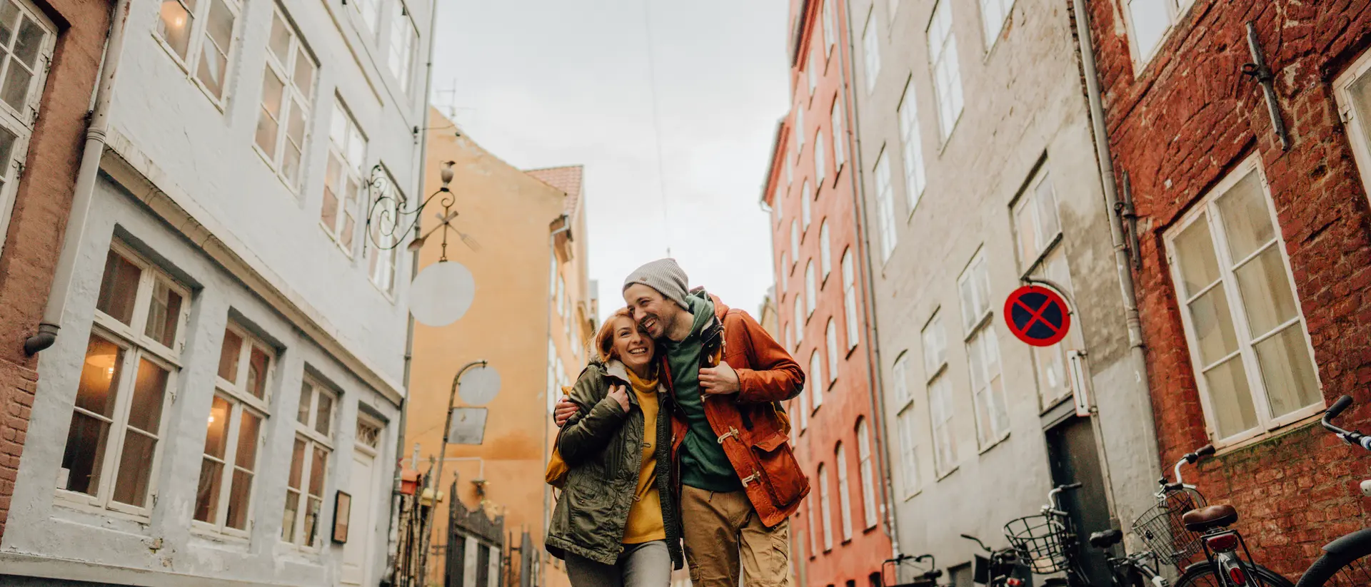 A happy couple stands on a cobbled street, surrounded by colorful buildings and bicycles parked on the side.