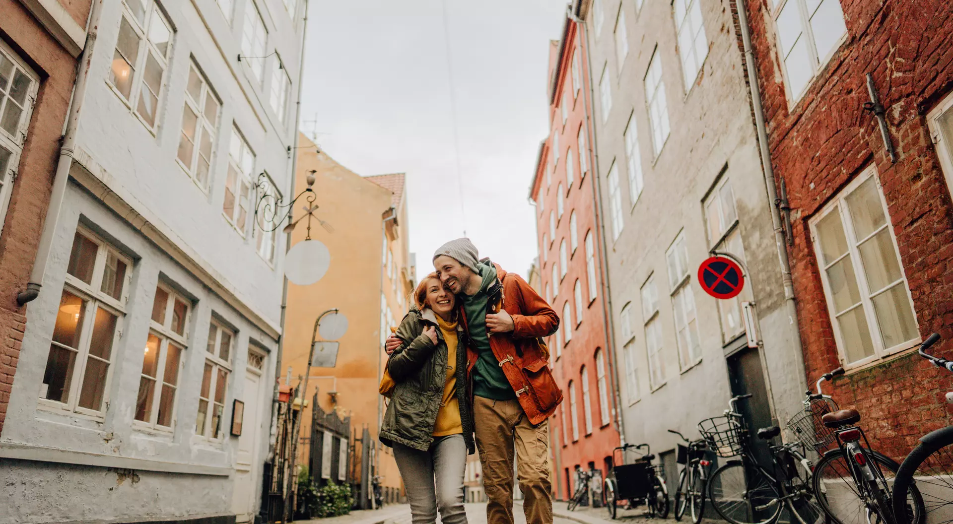 A happy couple stands on a cobbled street, surrounded by colorful buildings and bicycles parked on the side.