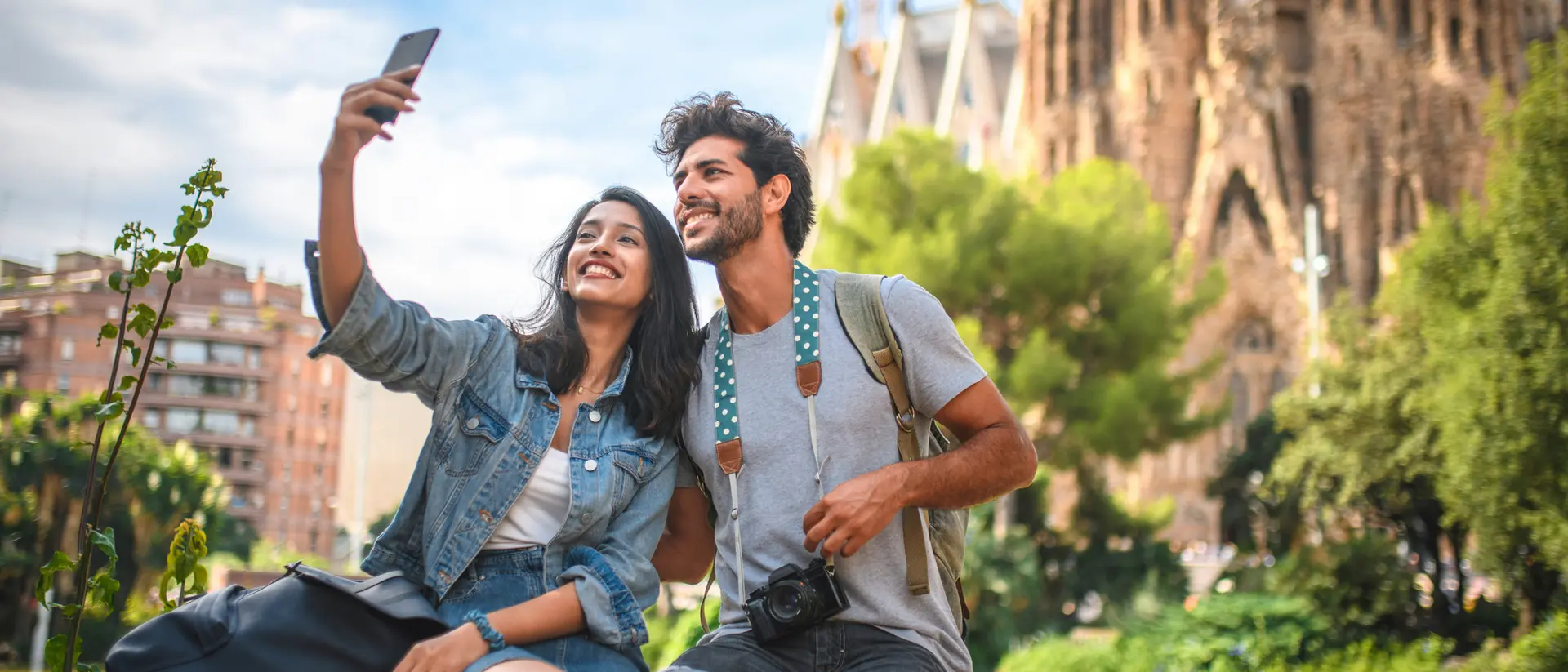 A smiling couple takes a selfie outdoors, with greenery and an iconic building in the background.