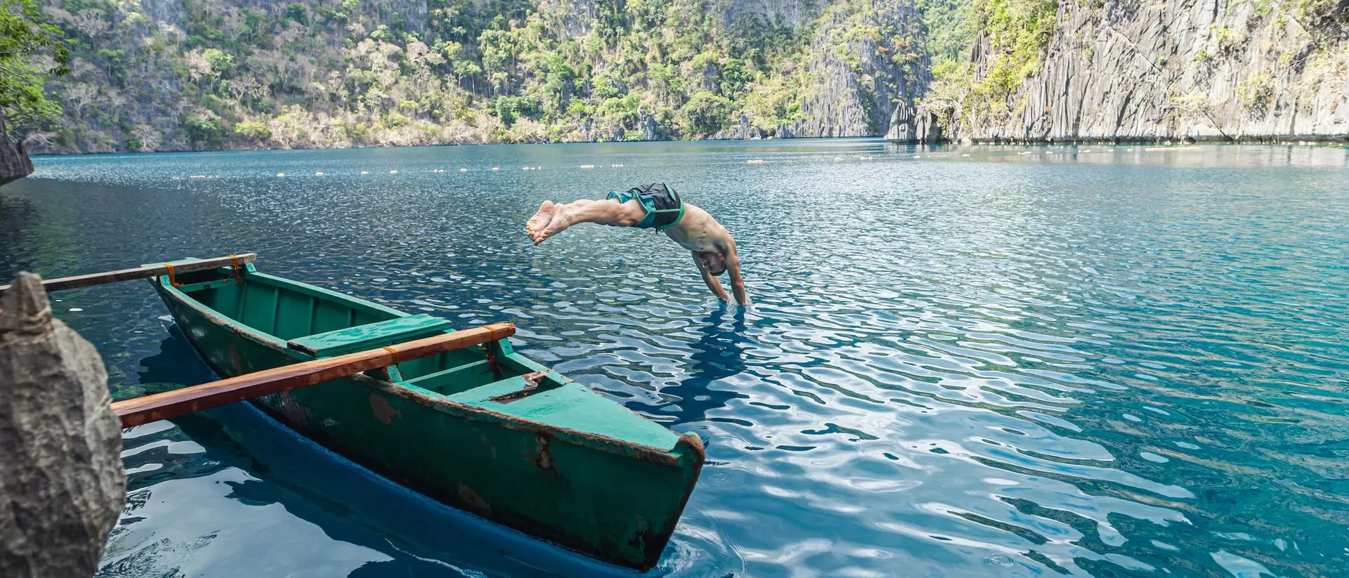 A person dives into clear blue water from the edge of a green boat surrounded by rocky cliffs and lush greenery.
