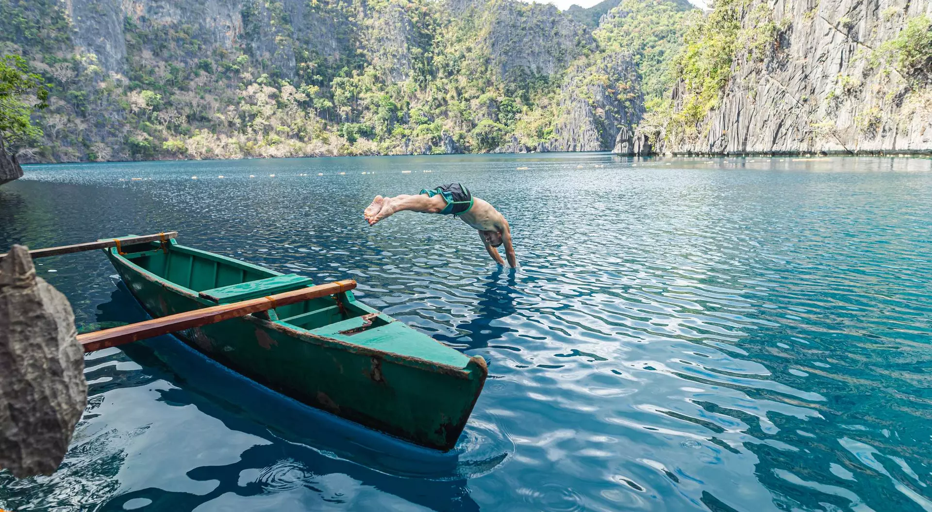 A person dives into clear blue water from the edge of a green boat surrounded by rocky cliffs and lush greenery.