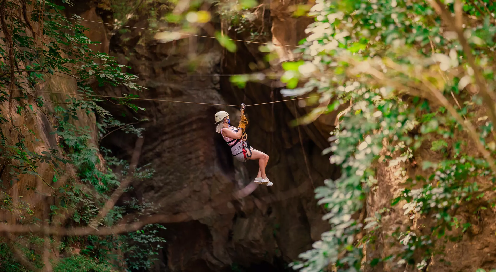 A person zip-lining through a lush, green forest with rocky cliffs in the background.