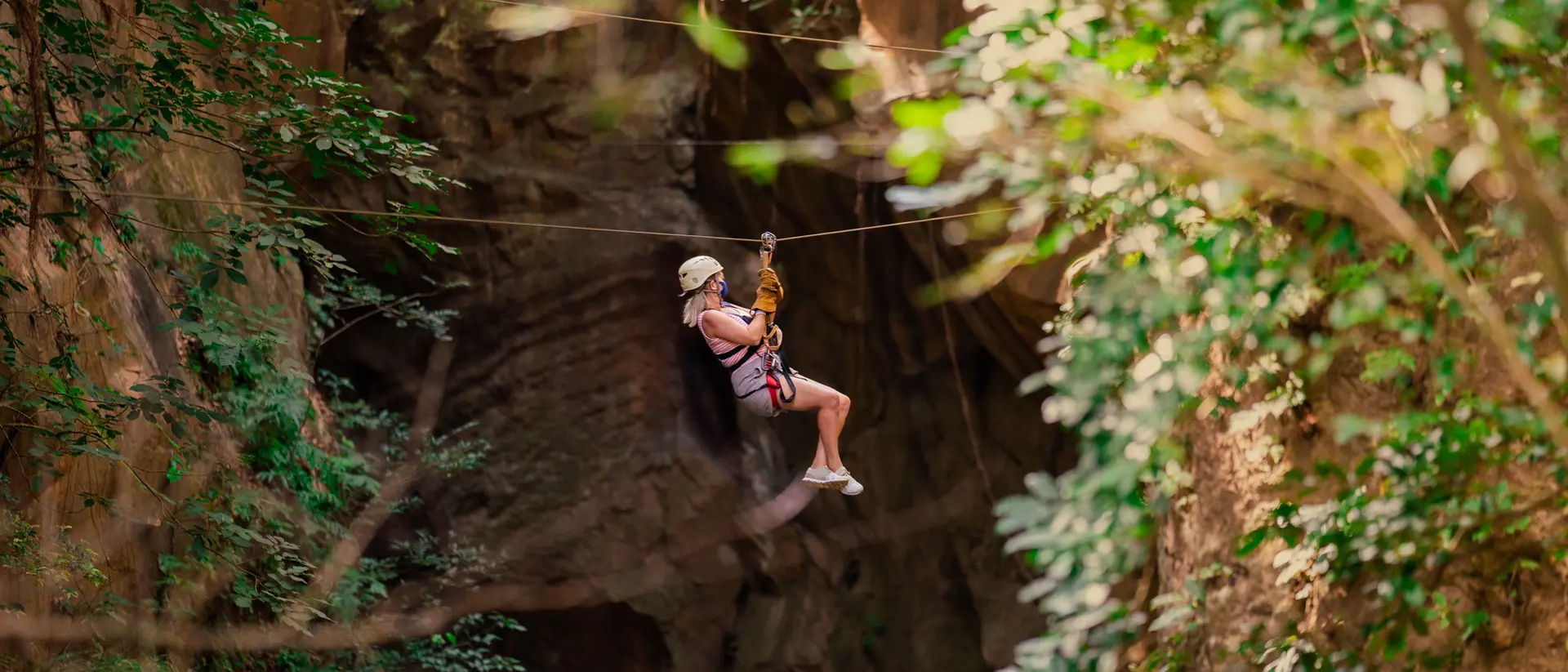 A person zip-lining through a lush, green forest with rocky cliffs in the background.