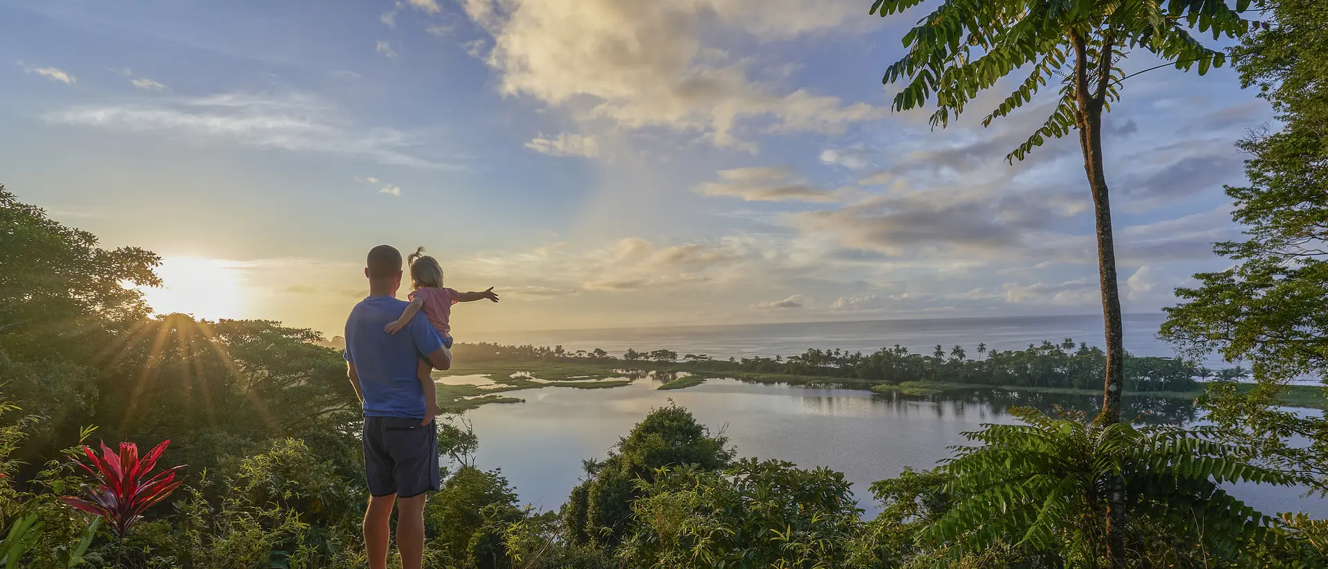 A parent holds a child while gazing at a scenic view of a lake and lush landscape under a bright sky.