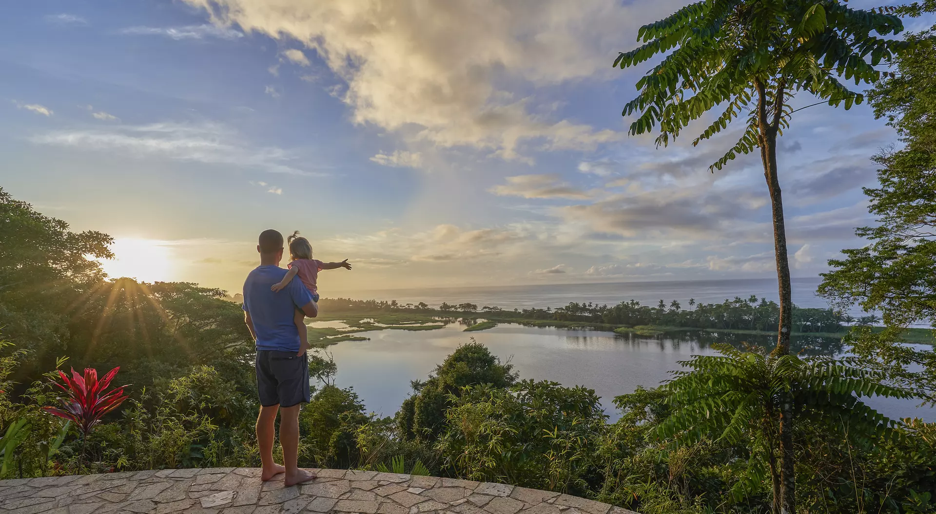 A parent holds a child while gazing at a scenic view of a lake and lush landscape under a bright sky.