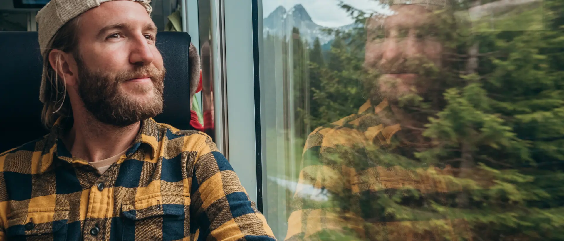 A man in a plaid shirt and cap gazes out the train window, with mountains and trees reflected in the glass.