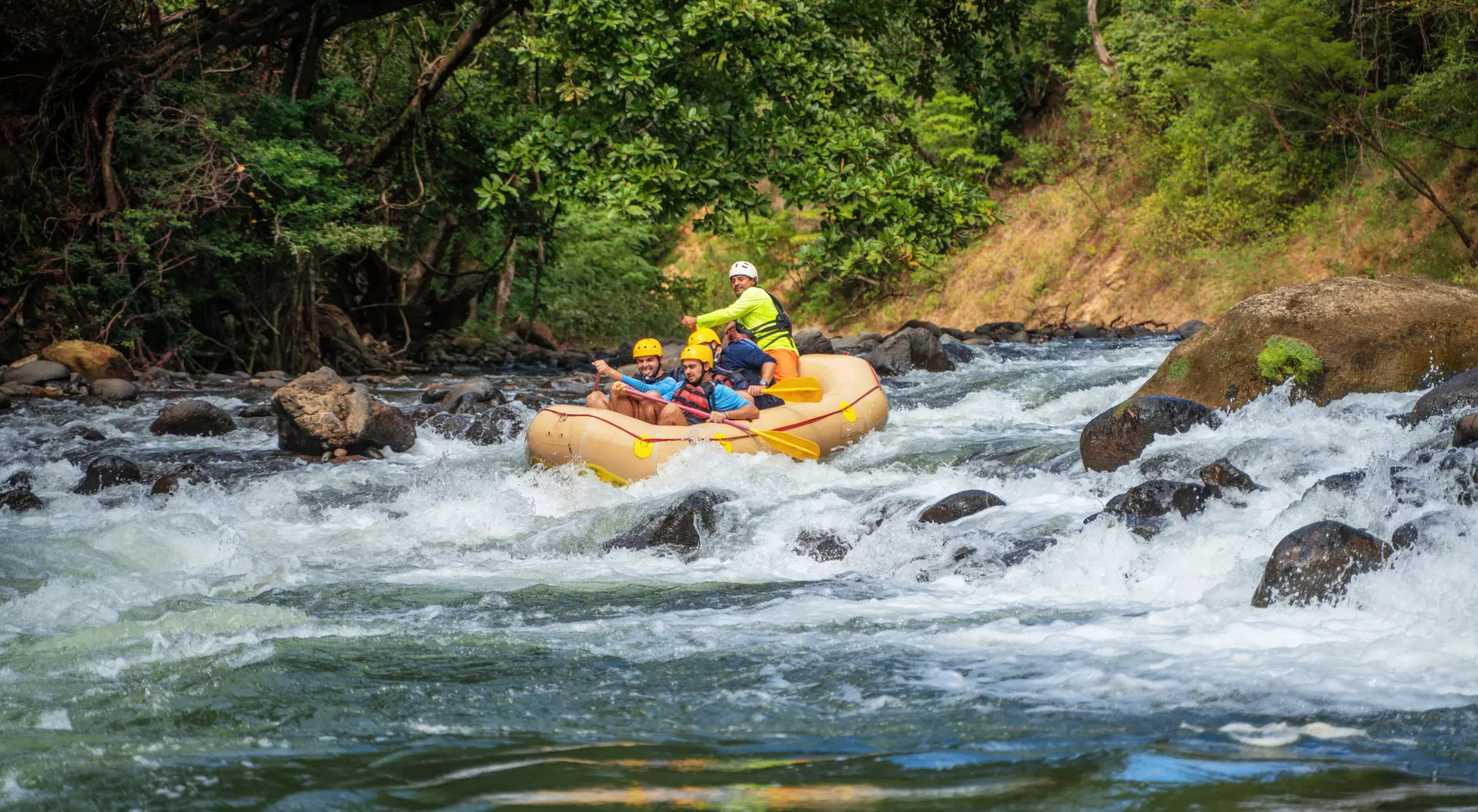 Two people navigate a yellow raft through turbulent river waters surrounded by lush green trees.