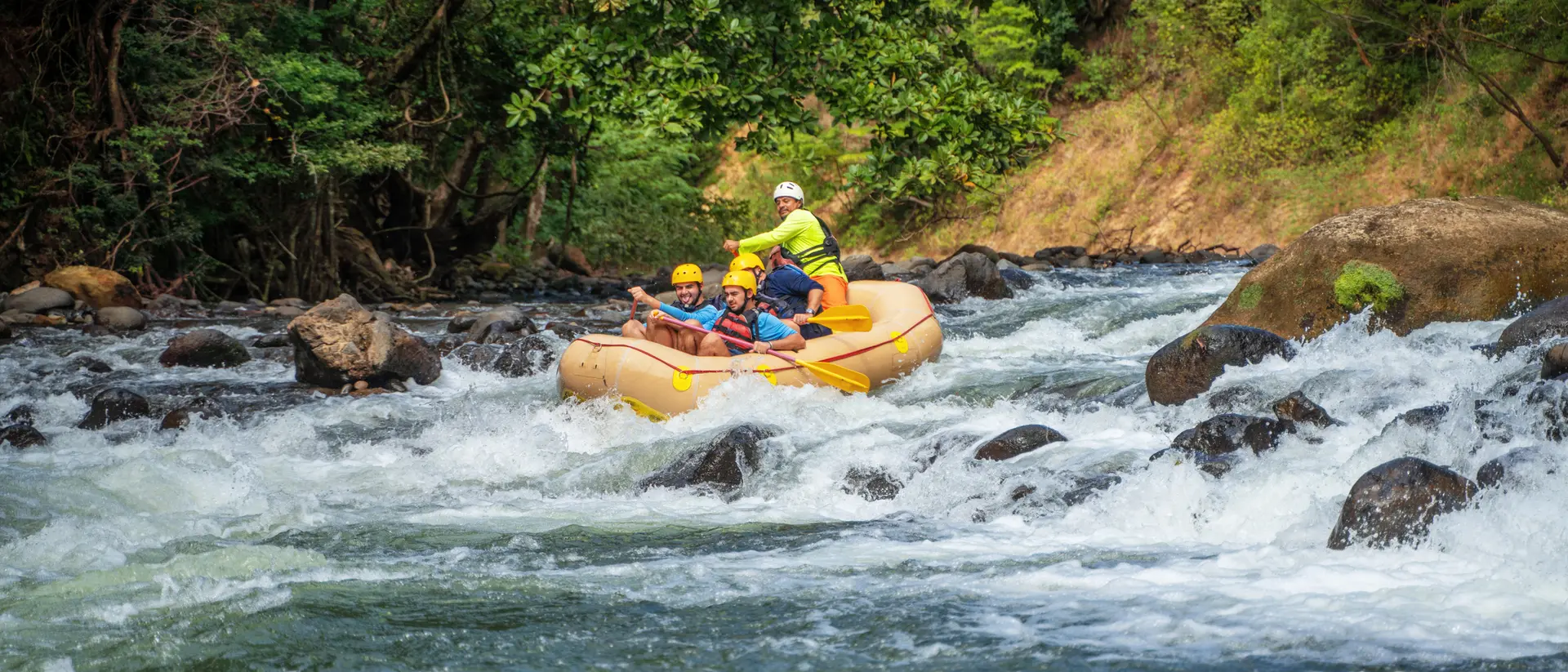 Two people navigate a yellow raft through turbulent river waters surrounded by lush green trees.