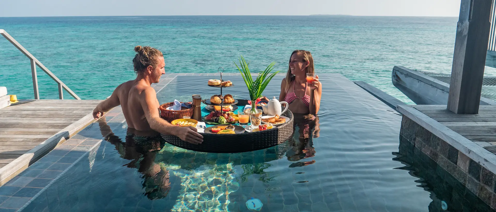 A couple relaxes in a pool, enjoying a floating breakfast with scenic ocean views.