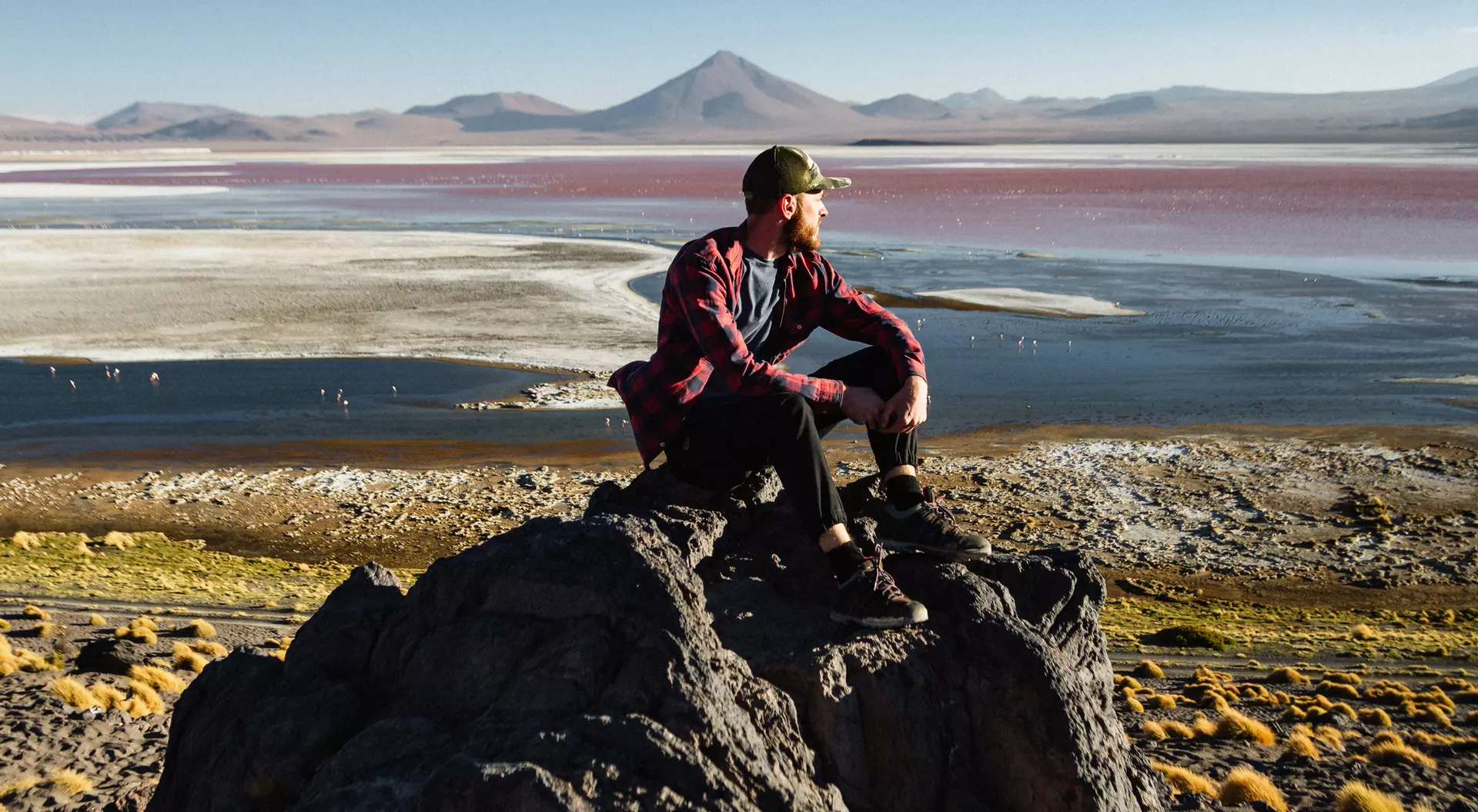 A person sits on a rock overlooking a colorful landscape with a body of water and distant mountains.