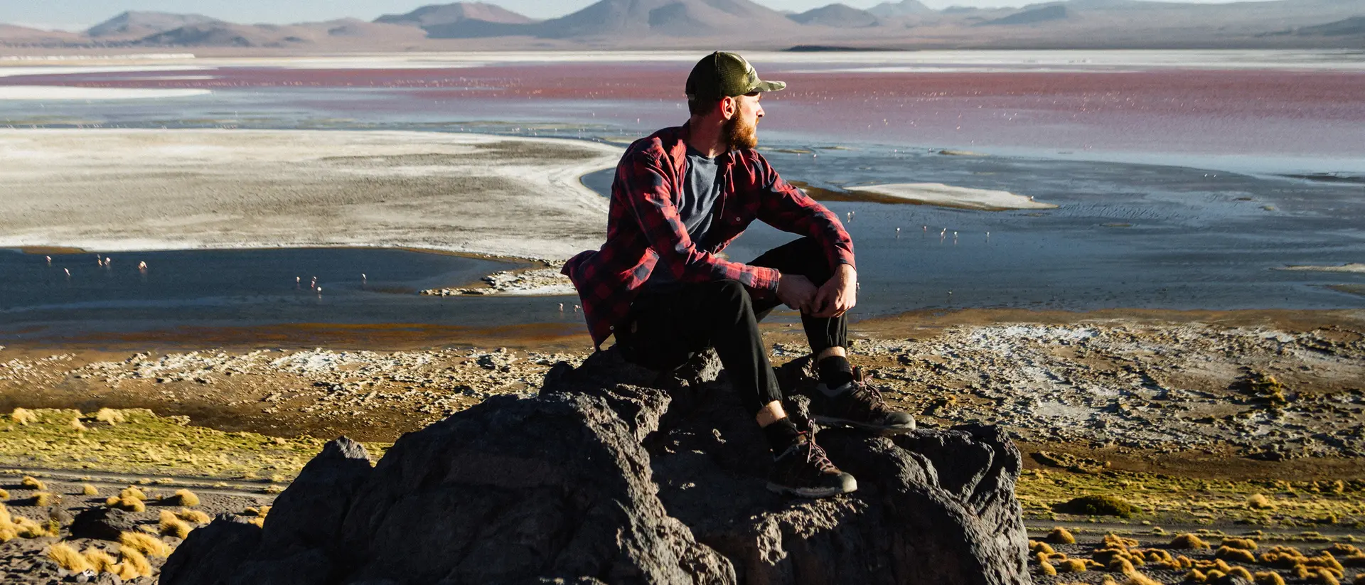 A person sits on a rock overlooking a colorful landscape with a body of water and distant mountains.