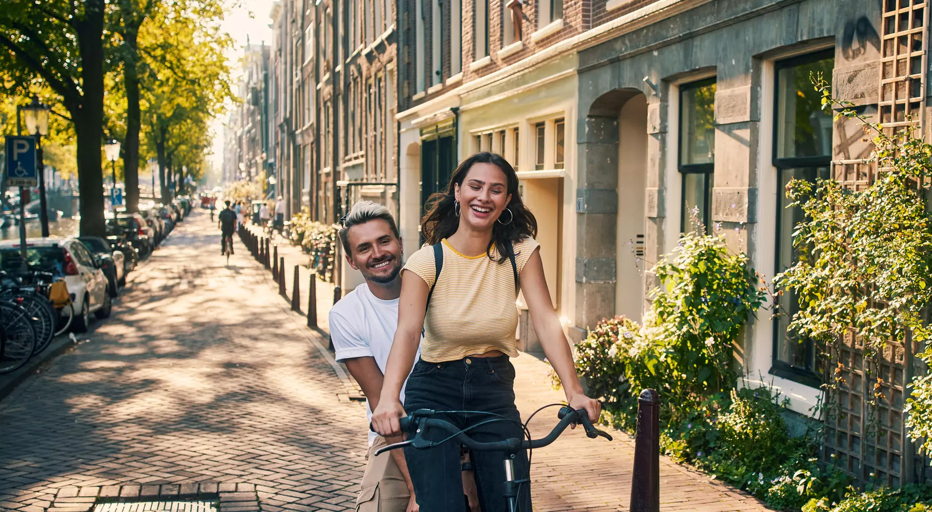 A woman smiles while riding a bike, with a man playfully behind her, on a sunny street lined with trees and buildings.