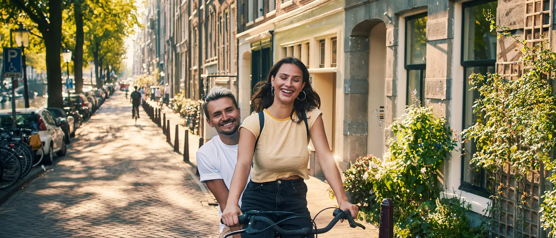 A woman smiles while riding a bike, with a man playfully behind her, on a sunny street lined with trees and buildings.