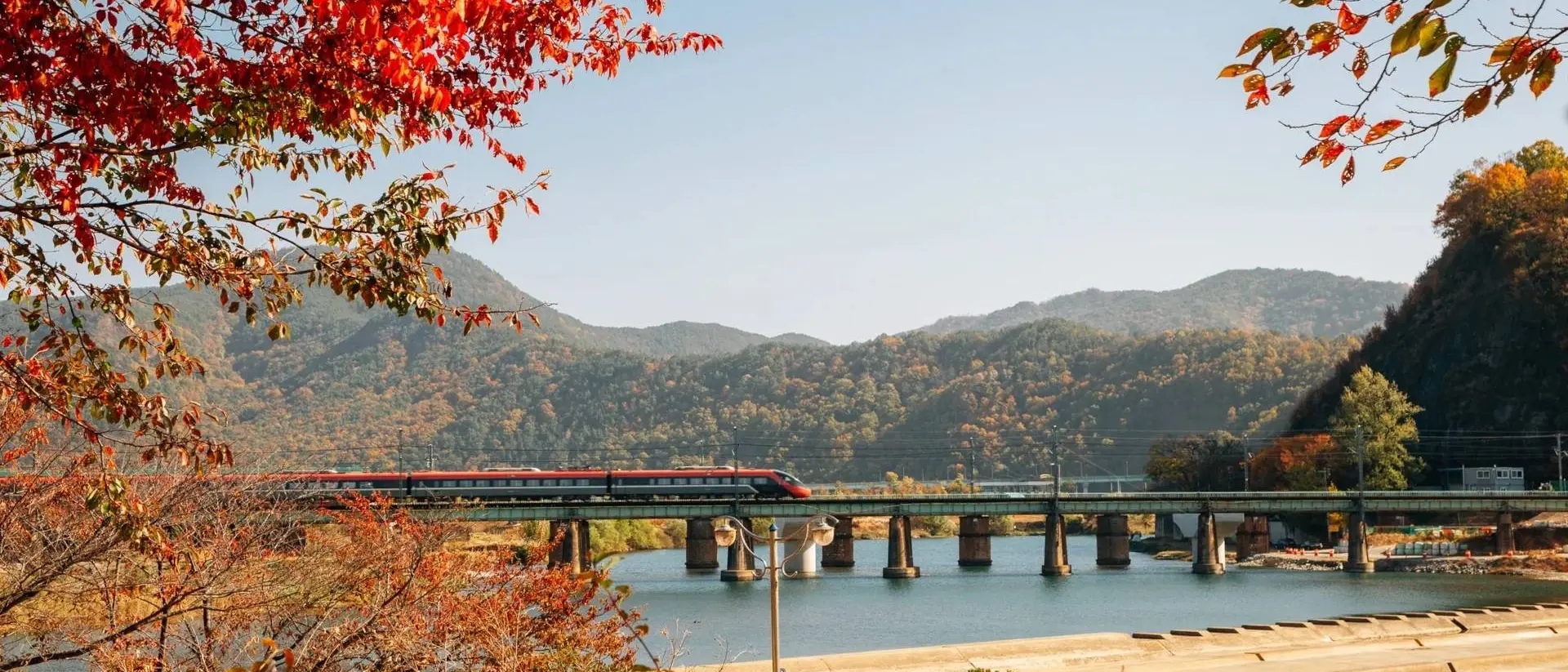 A train crosses a bridge over a river, surrounded by autumn foliage and mountains under a clear blue sky.