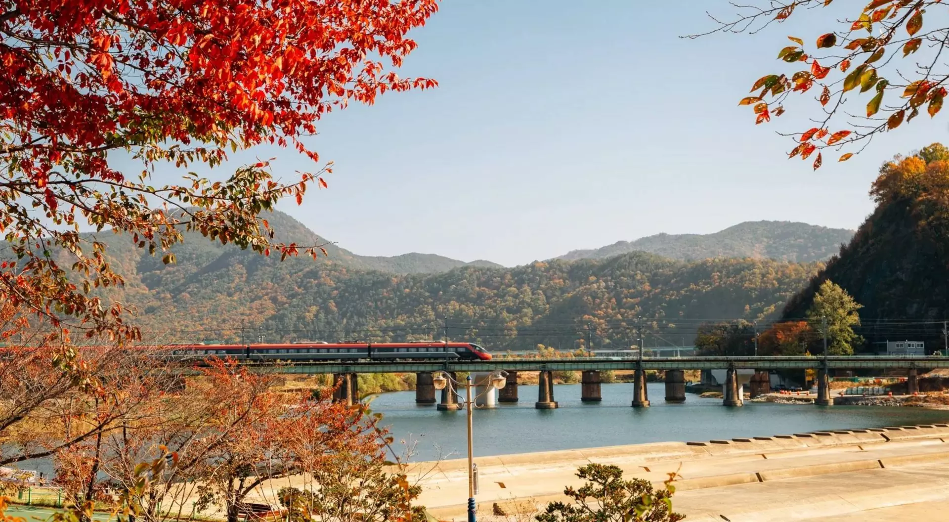 A train crosses a bridge over a river, surrounded by autumn foliage and mountains under a clear blue sky.