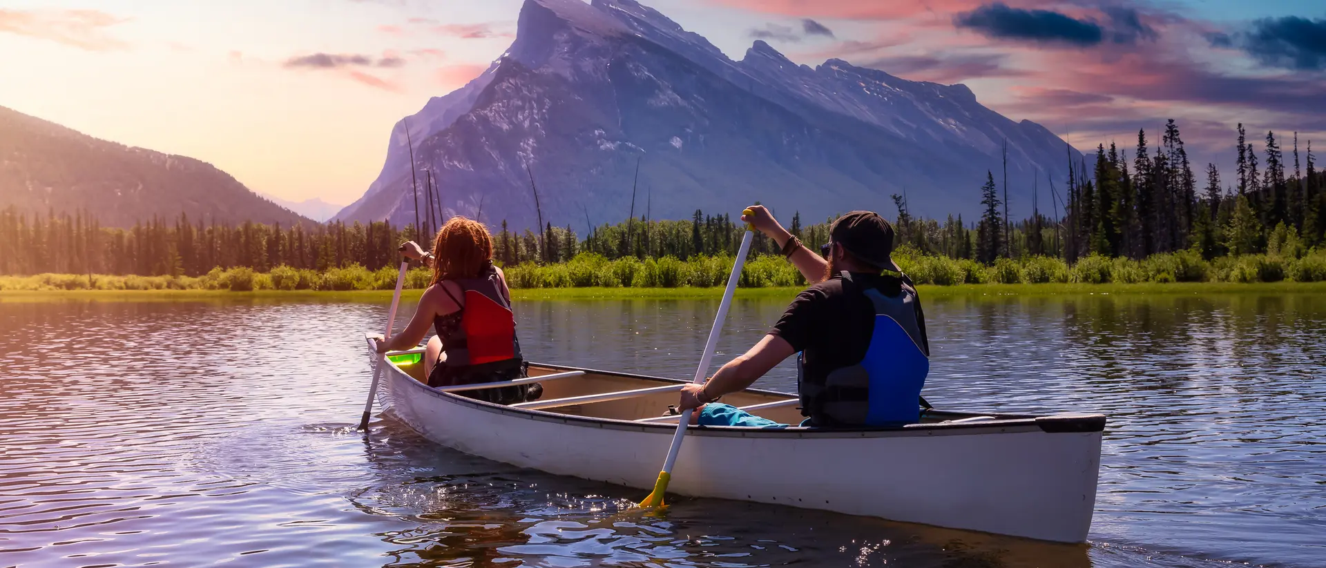 Two people paddle a canoe on a calm lake, with mountains and colorful sunset in the background.