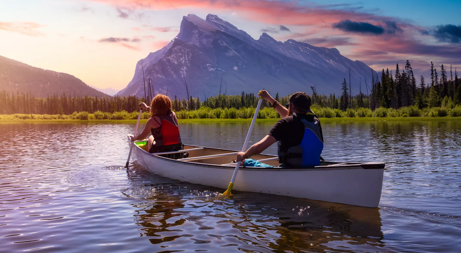 Two people paddle a canoe on a calm lake, with mountains and colorful sunset in the background.