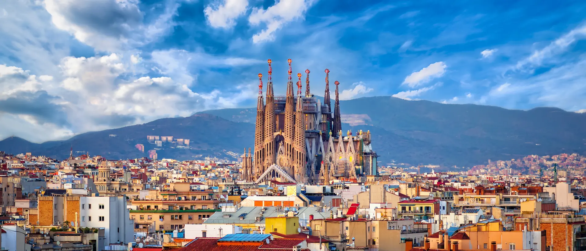 Panoramic view of Barcelona featuring the Sagrada Família, surrounded by city rooftops and a bright blue sky.