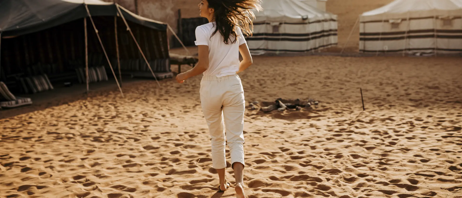 A woman in a white outfit walks barefoot through sand, heading towards tents in a desert landscape.