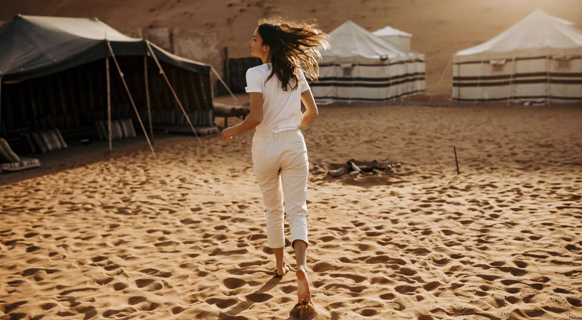 A woman in a white outfit walks barefoot through sand, heading towards tents in a desert landscape.
