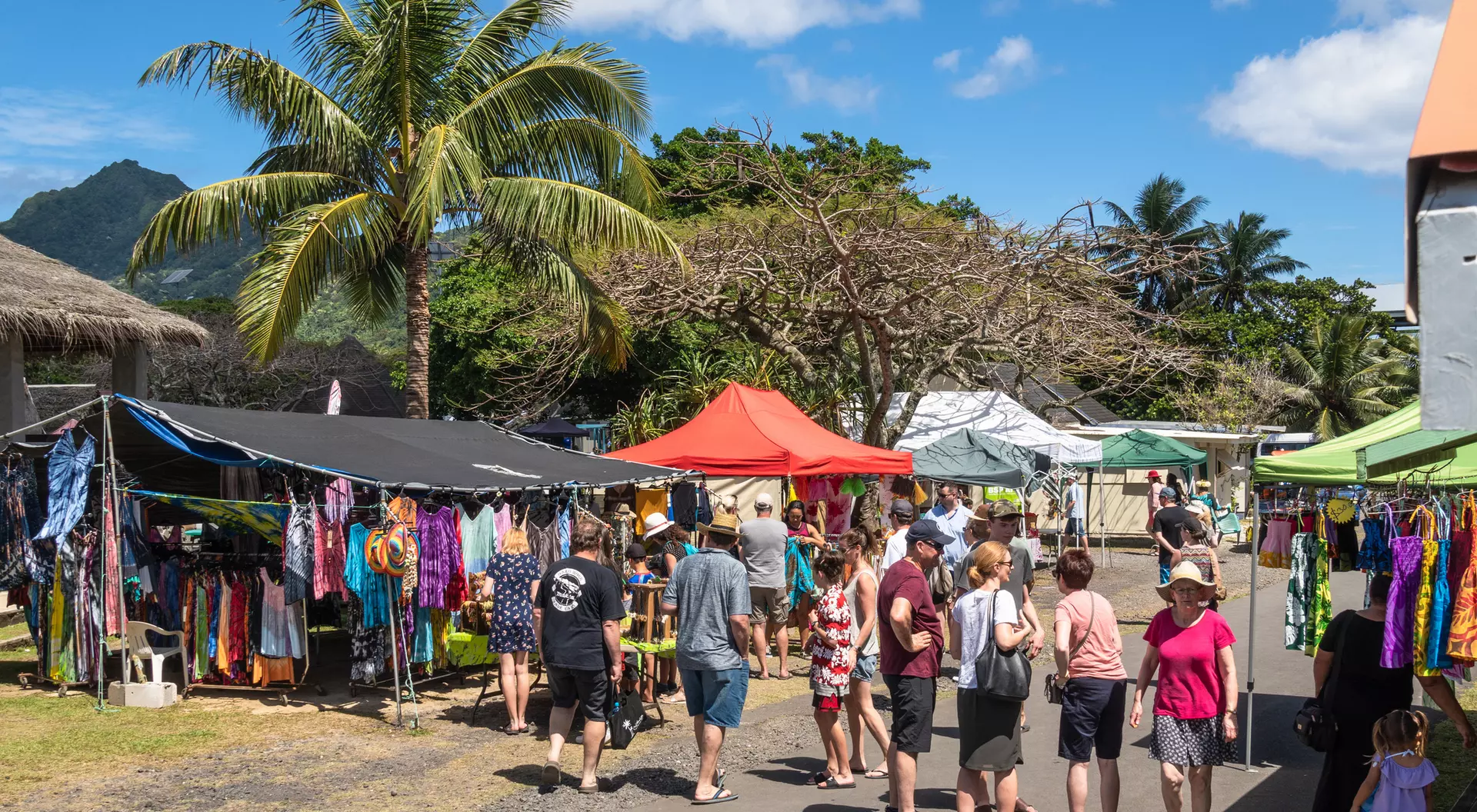 A vibrant outdoor market scene with tents selling clothing, surrounded by palm trees and a crowd of shoppers enjoying the day.