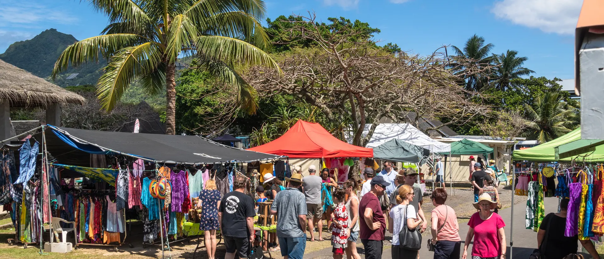 A vibrant outdoor market scene with tents selling clothing, surrounded by palm trees and a crowd of shoppers enjoying the day.