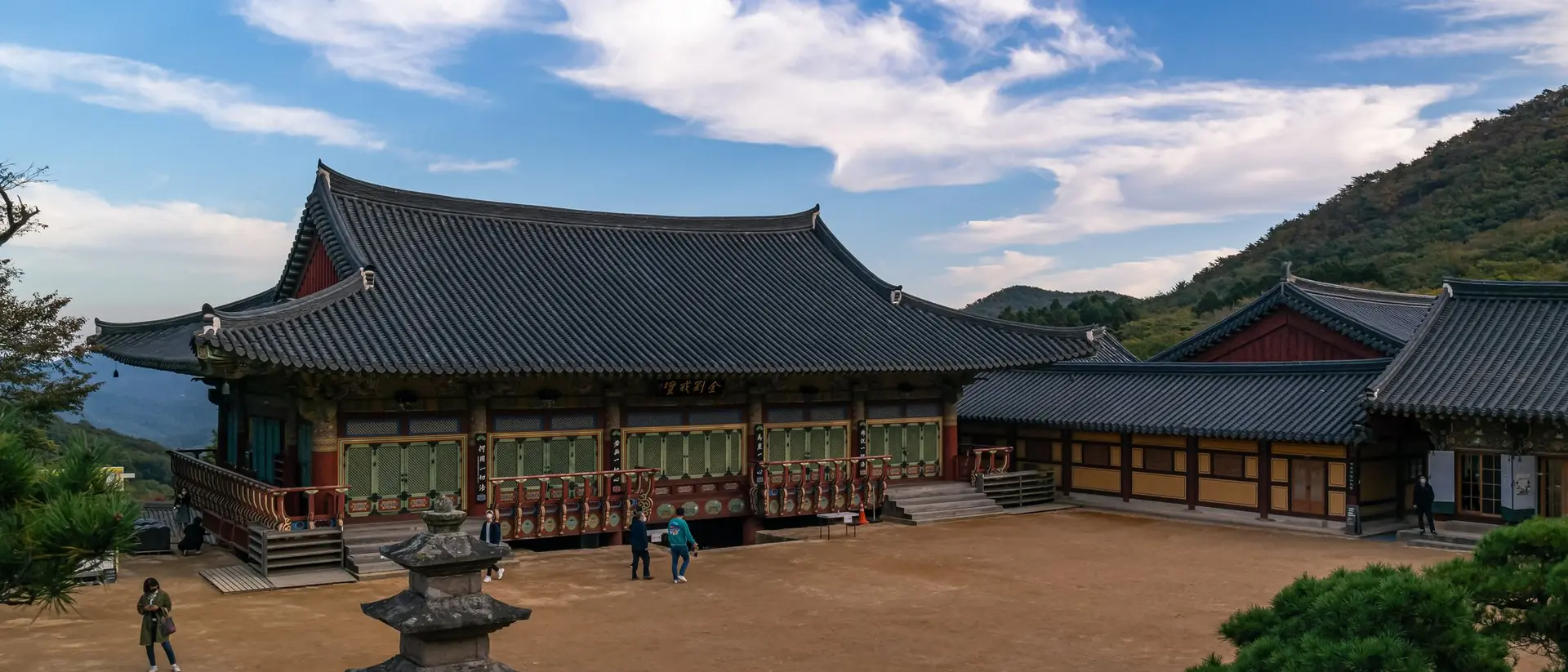 Historic temple with traditional Korean architecture, surrounded by mountains and a clear blue sky. Visitors stroll the grounds.