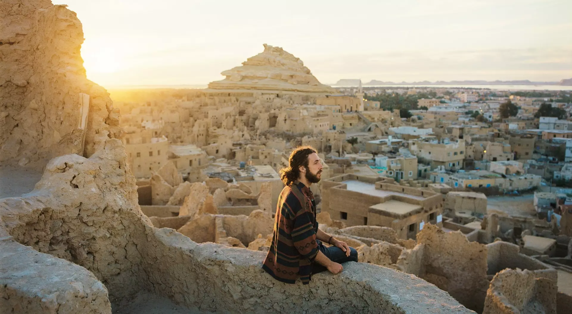 A person sits on a wall overlooking a village at sunset, with ancient structures and a pyramid in the background.