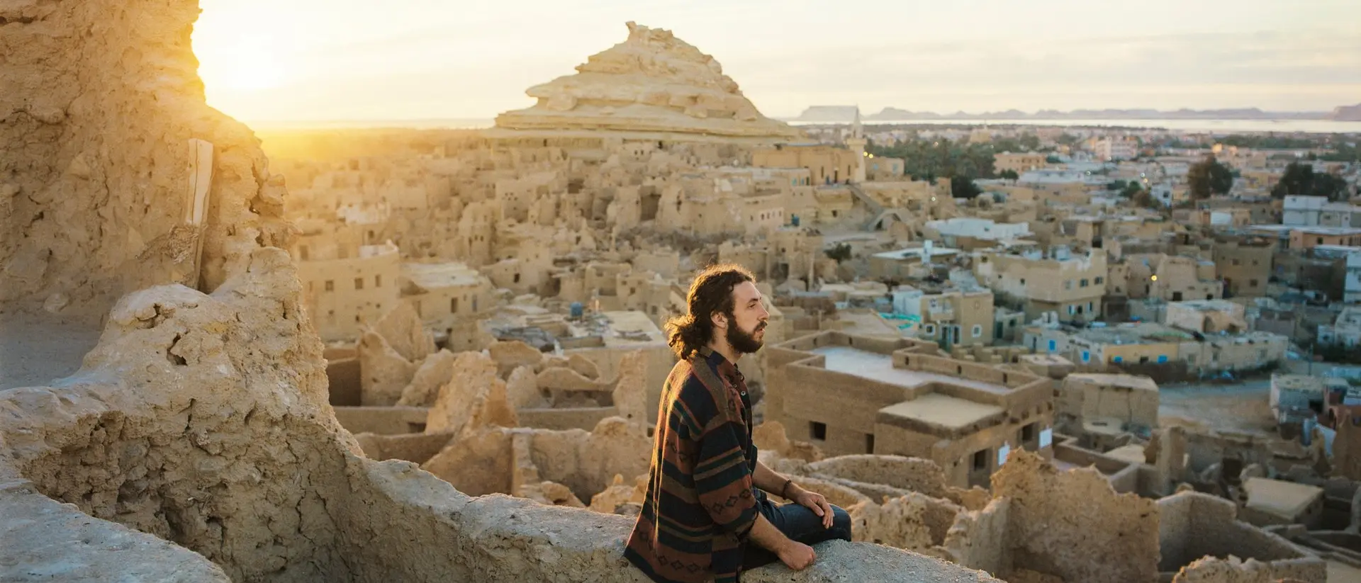 A person sits on a wall overlooking a village at sunset, with ancient structures and a pyramid in the background.