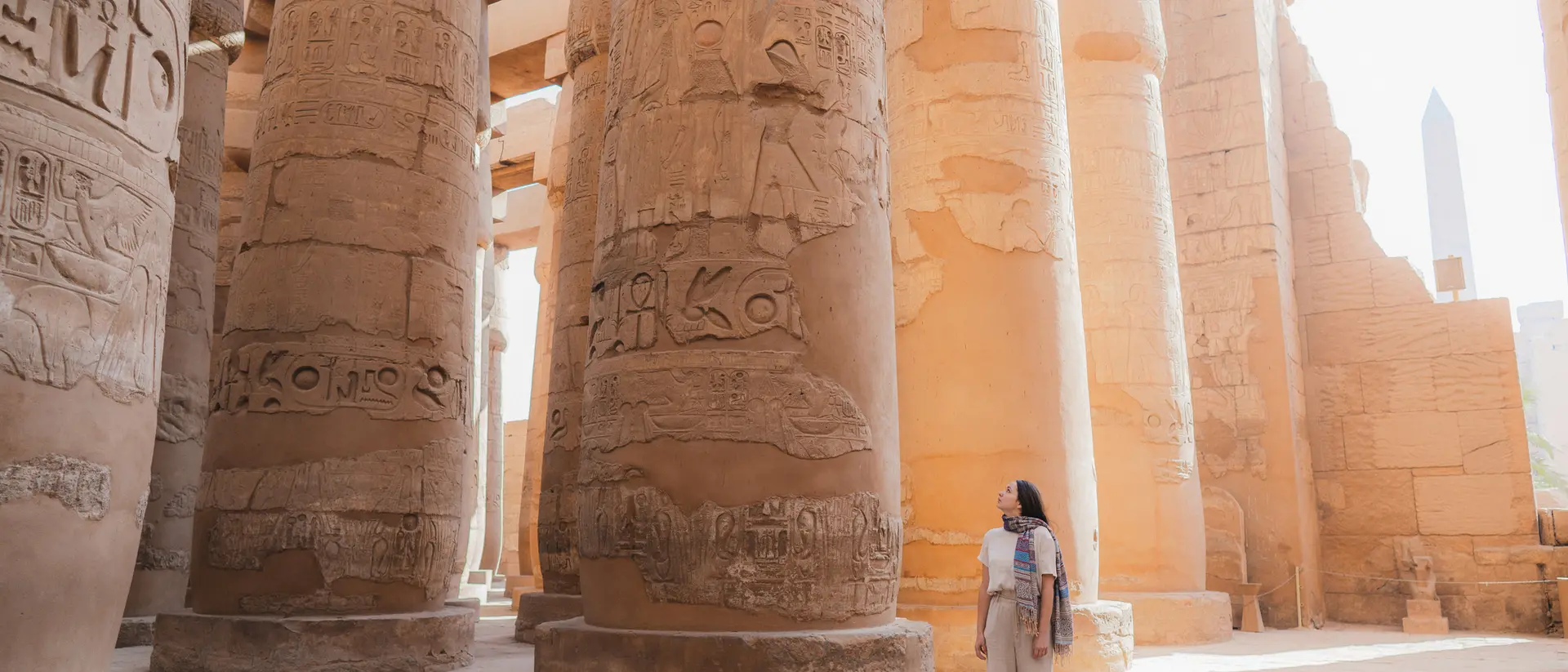 Person standing among tall, ancient, carved stone columns inside a historic site, with sunlight streaming in.