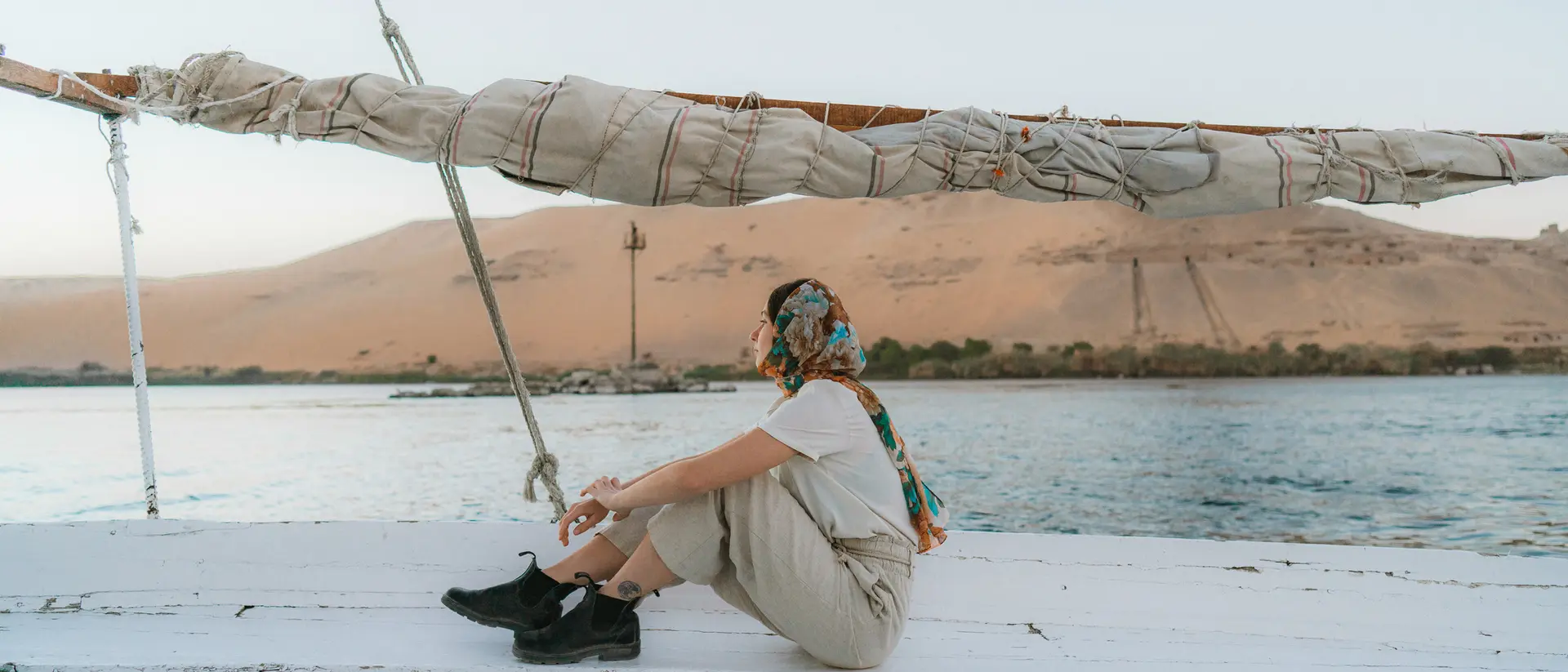 Person sitting on a boat, wearing a scarf, with desert hills and water in the background during sunset.
