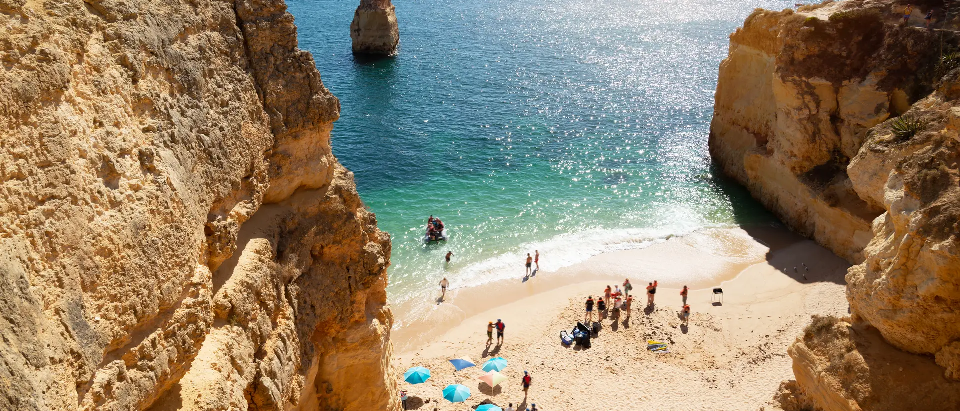 A sunlit beach with people near turquoise water, surrounded by rocky cliffs and colorful umbrellas.