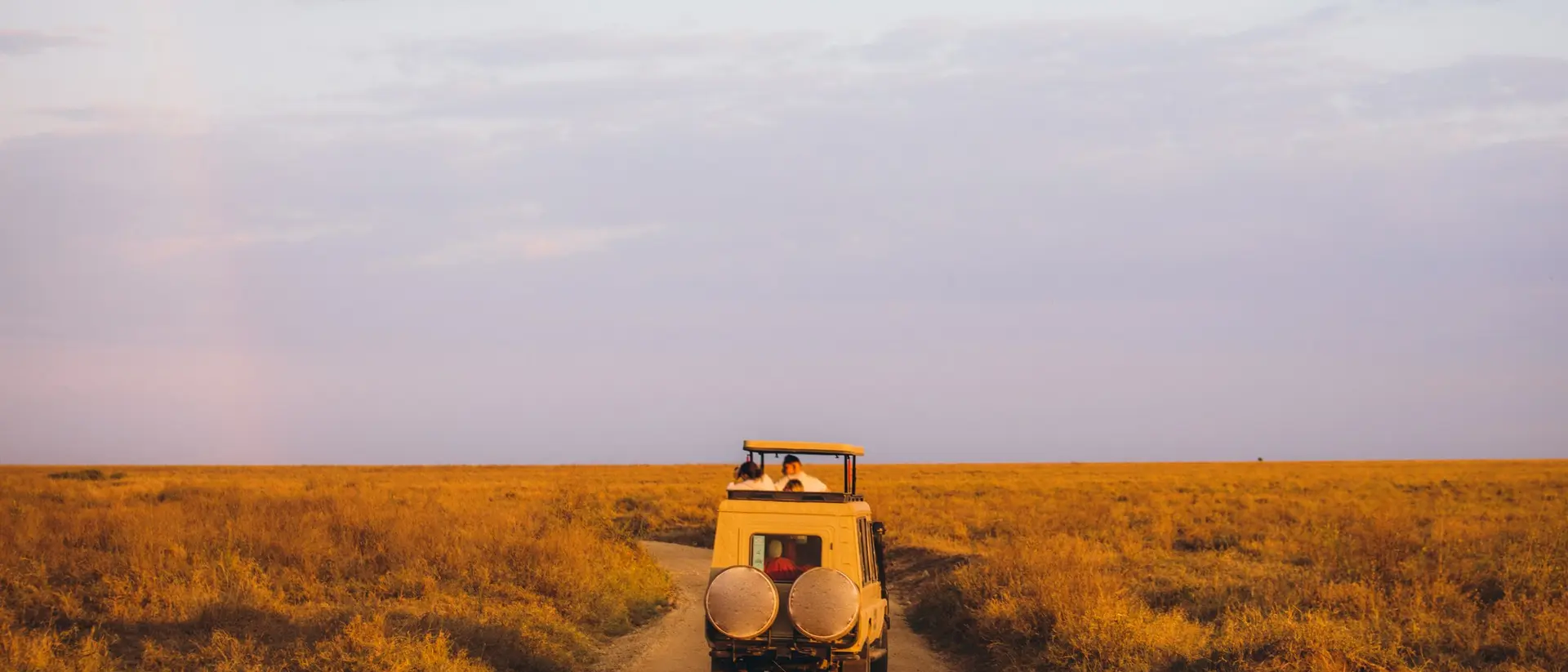 A safari vehicle drives along a dirt road in an open grassland at sunset, with a colorful sky overhead.