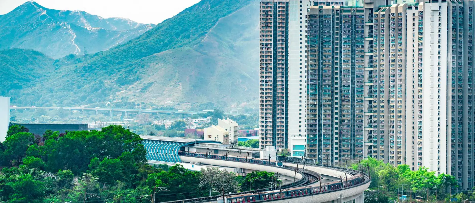 A train travels along a curved track near tall buildings and lush green trees, with mountains in the background.