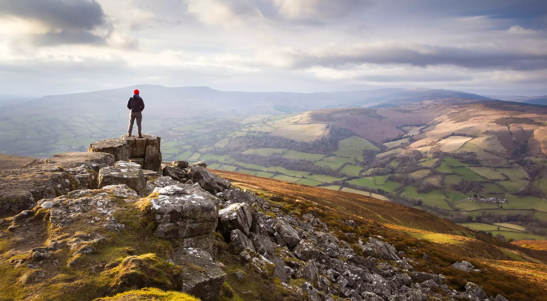 A person stands on a rocky outcrop, overlooking a scenic valley with rolling hills and a cloudy sky.