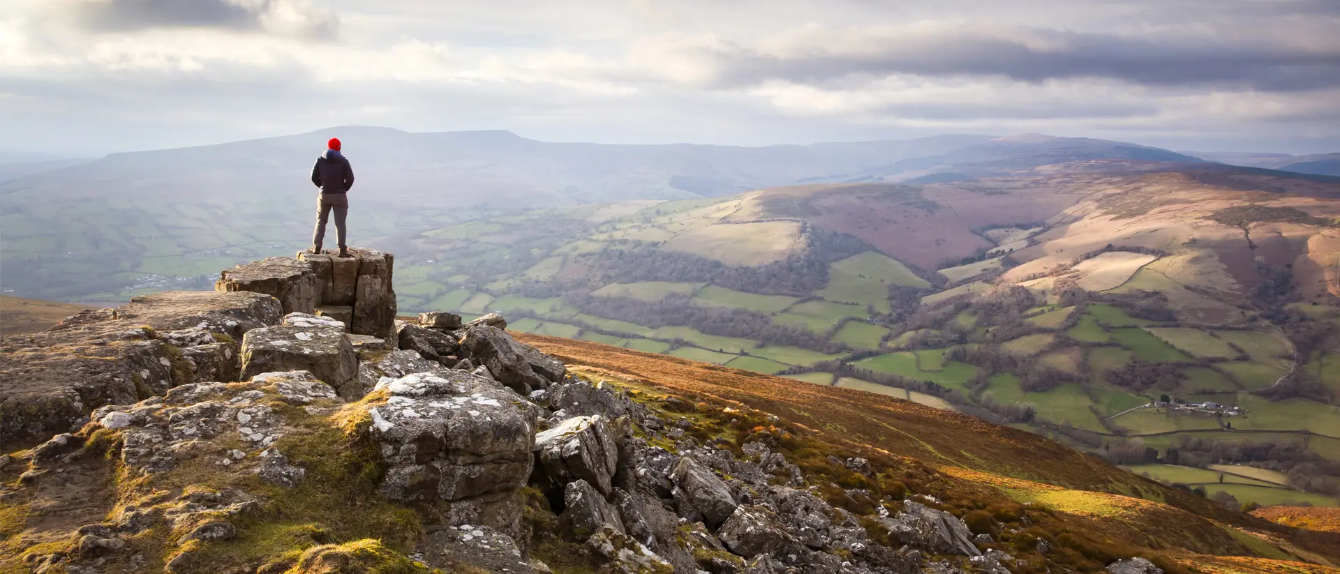 A person stands on a rocky outcrop, overlooking a scenic valley with rolling hills and a cloudy sky.
