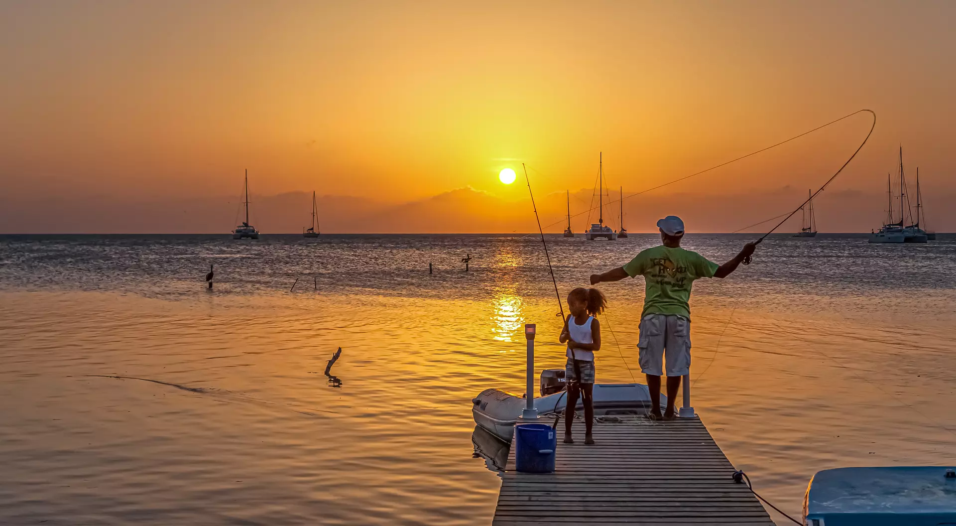 A man and a child fish from a dock at sunset, with boats in the background and calm water reflecting the orange sky.