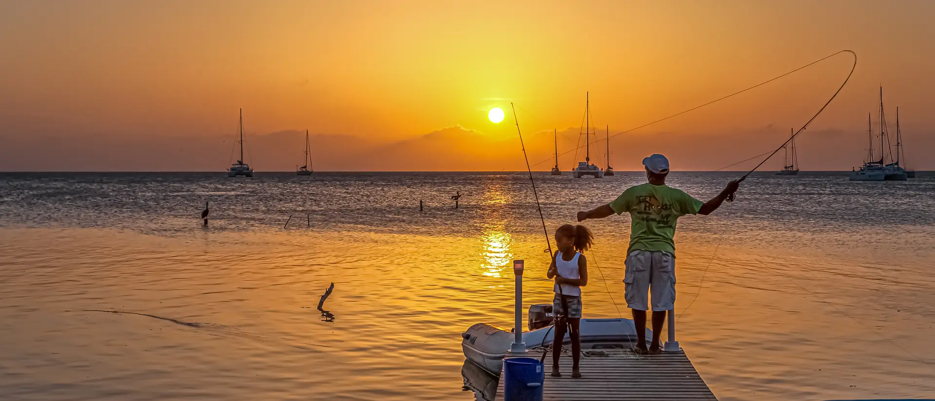 A man and a child fish from a dock at sunset, with boats in the background and calm water reflecting the orange sky.