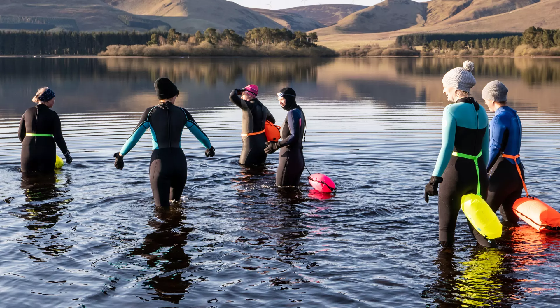 A group of six people in wetsuits entering a calm lake surrounded by hills on a sunny day.