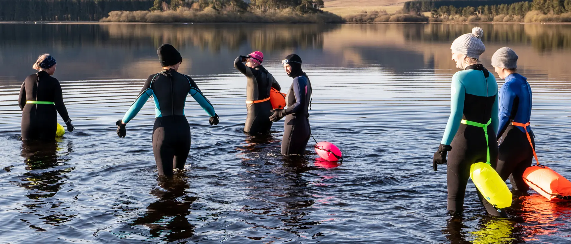 A group of six people in wetsuits entering a calm lake surrounded by hills on a sunny day.