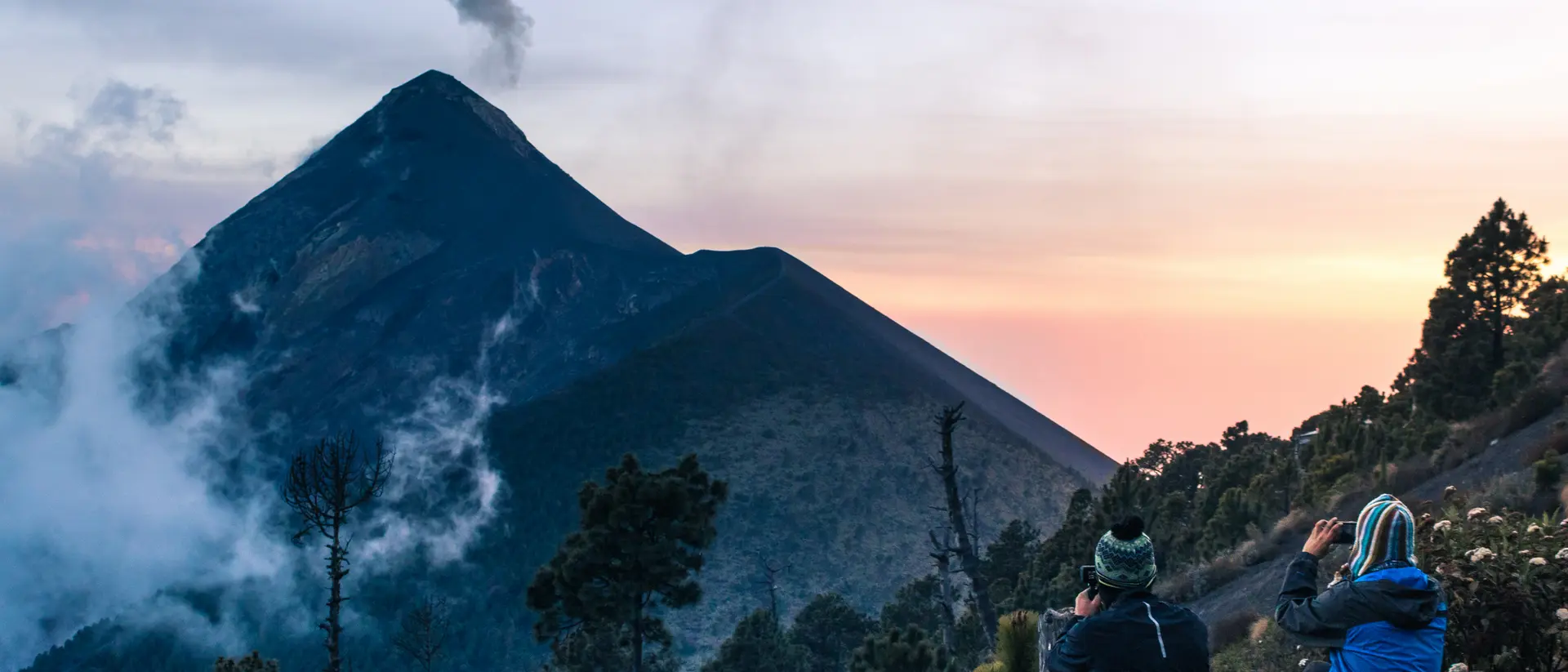 Two hikers sit on a rocky ledge, taking photos of a tall volcano against a colorful sunset sky.