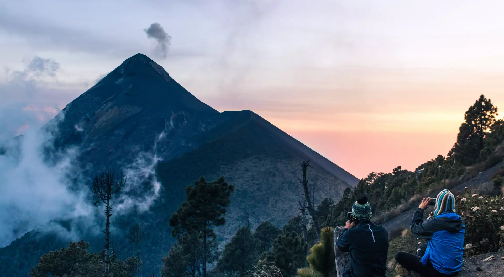 Two hikers sit on a rocky ledge, taking photos of a tall volcano against a colorful sunset sky.