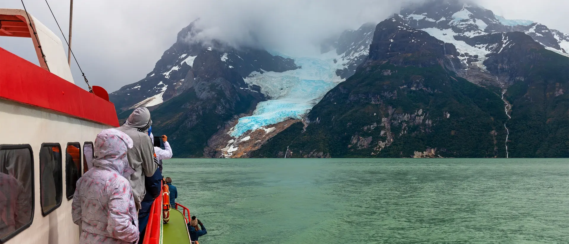Passengers on a boat view a glacier and mountains under a cloudy sky, with water visible in the foreground.
