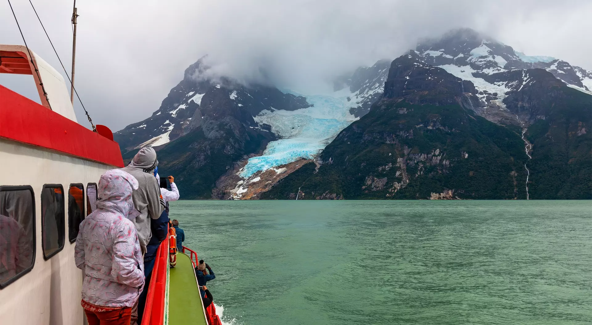 Passengers on a boat view a glacier and mountains under a cloudy sky, with water visible in the foreground.