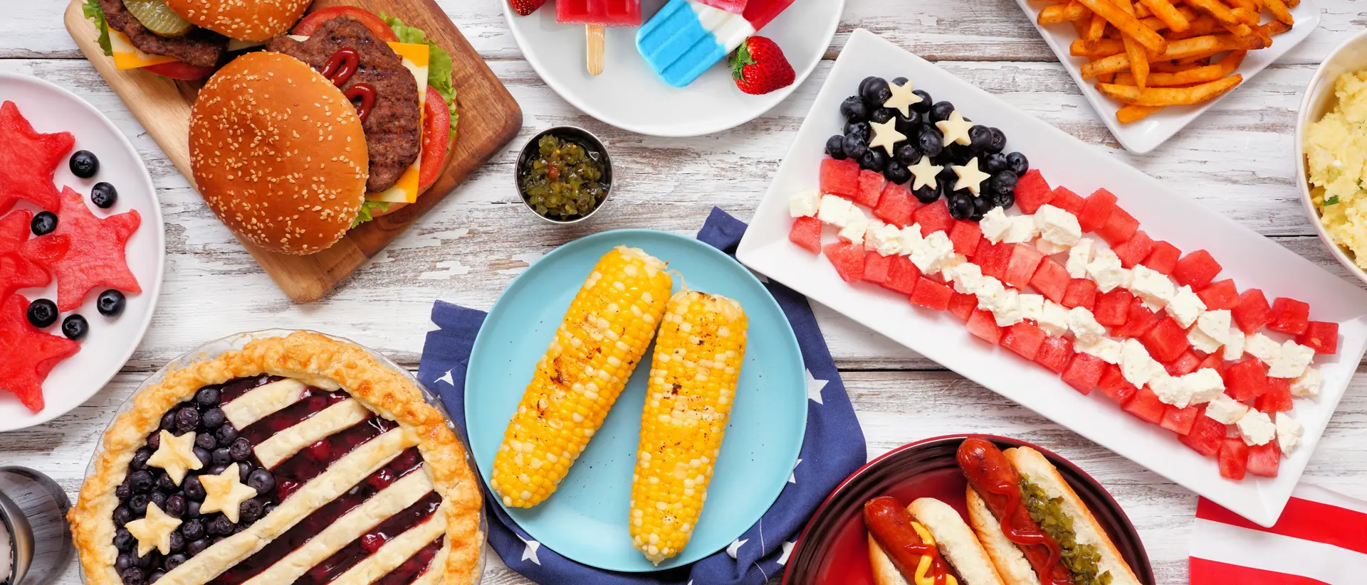 A variety of festive foods including burgers, hot dogs, watermelon, corn, and desserts arranged in a patriotic theme.