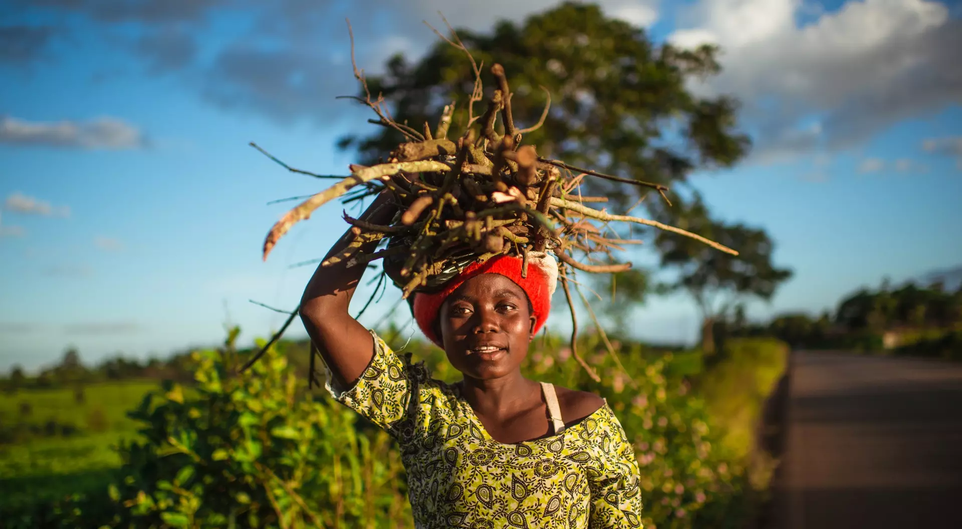 A girl with a red headwrap carries a large bundle of sticks on her head, standing on a rural road with greenery around.