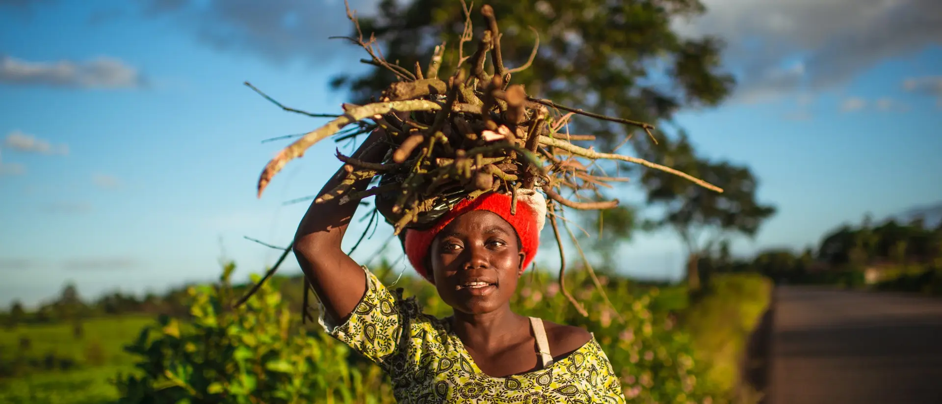 A girl with a red headwrap carries a large bundle of sticks on her head, standing on a rural road with greenery around.