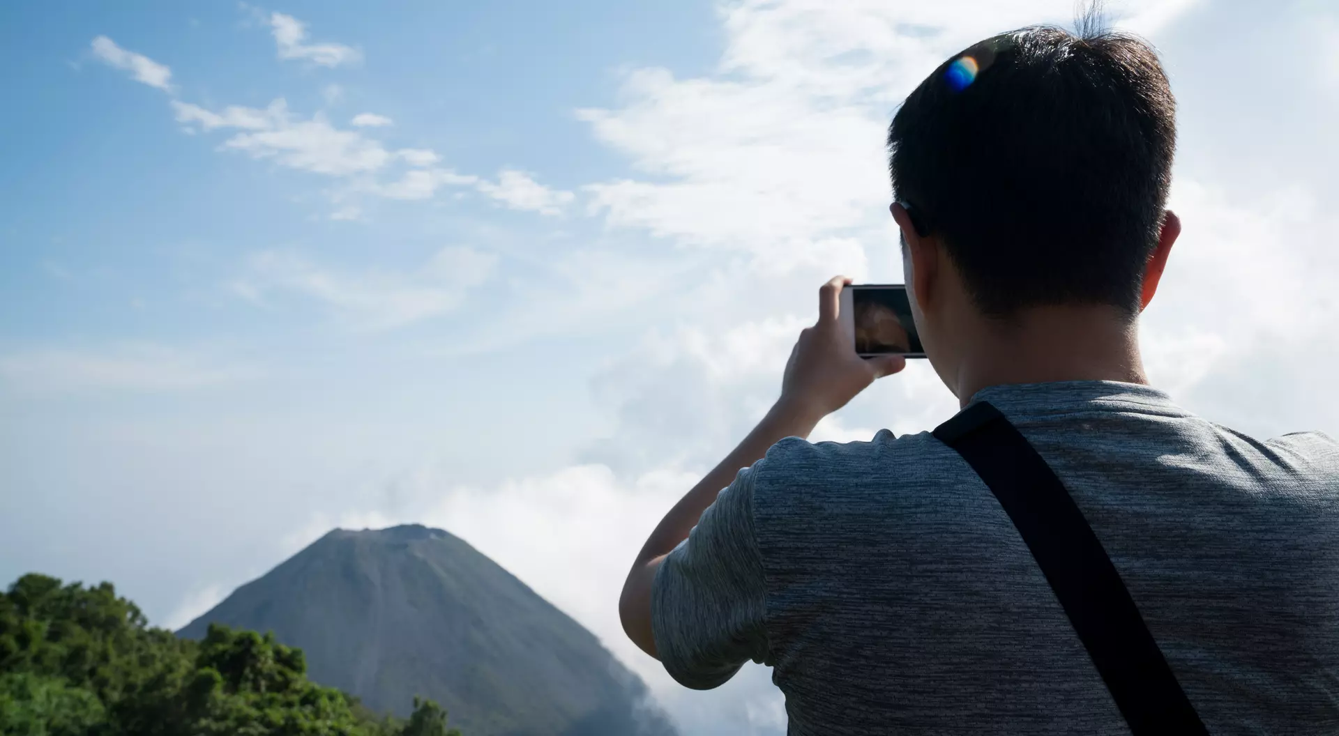 Person taking a photo of a volcano amidst greenery and clouds under a blue sky.