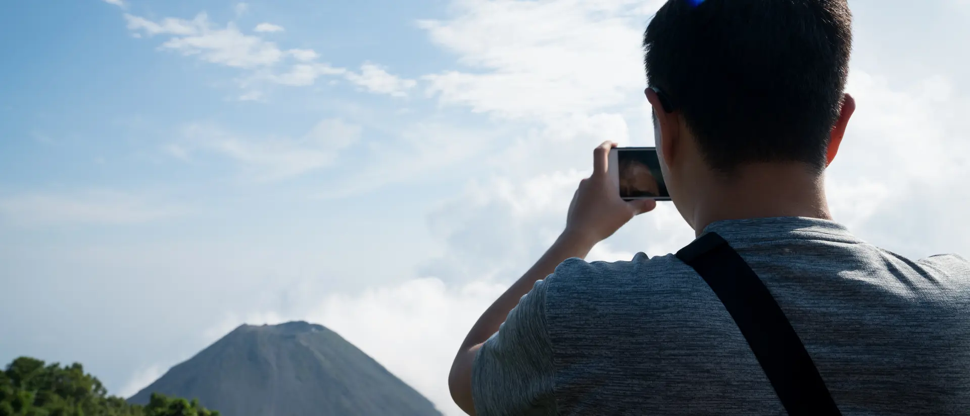 Person taking a photo of a volcano amidst greenery and clouds under a blue sky.
